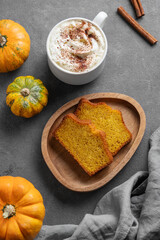 Slices of pumpkin sweet bread with cinnamon on a wooden board on a dark background with decorative pumpkins and a cup of hot chocolate or coffee.