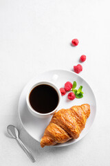 Cup of espresso coffee and croissant with fresh raspberries on a plate on a white texture  background with morning light.