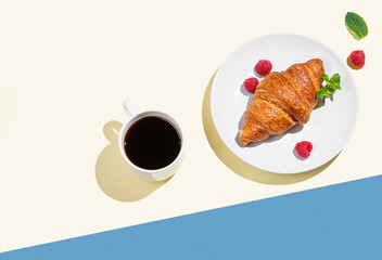 Fresh croissant on a white plate with raspberry and cup of espresso coffee on yellow and blue background.