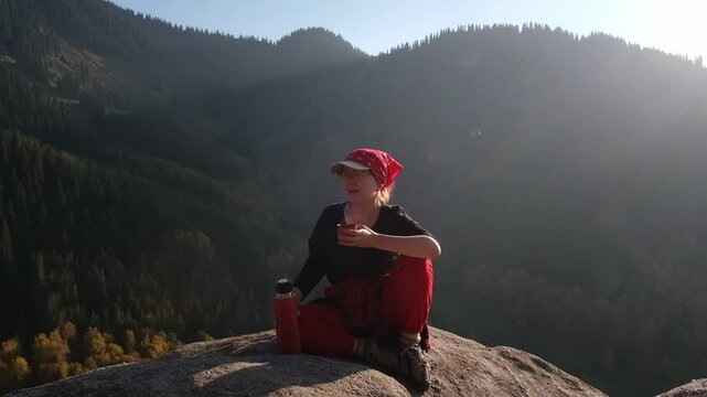 A woman enjoys a hot drink of tea or coffee from a red thermos against the backdrop of a beautiful landscape of autumn mountains.