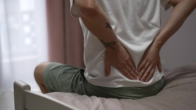 A man in a shirt sits on the bed and holds his lower back.
A guy has lower back pain.
The man's back is bothering him.
Back injury and severe lower back pain.
Trauma, pain, back, lower back, spine