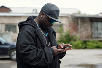 African American man wearing hat and sunglasses, engrossed in smartphone, standing in urban...