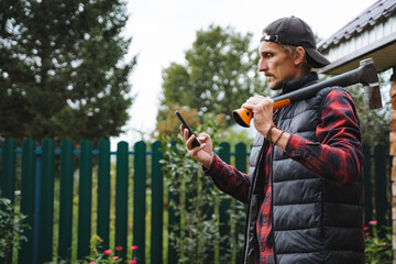 A person wearing a plaid shirt and a vest is using their phone while holding a hammer in a beautiful garden setting surrounded by greenery