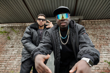 Two Black men wearing stylish street fashion with chains and sunglasses posing in front of brick...