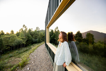 A woman leans against a modern cabin, reflecting on the tranquility of nature surrounding her. The...