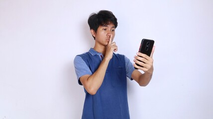 Asian young man gestures to shut up to close his mouth and holds smartphone