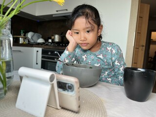 Asian little girl watching mobile phone after eating in the modern kitchen. Screen time health risks concept