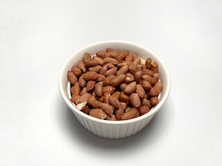 A white ceramic bowl filled with a heap of roasted peanuts. The peanuts are a light brown color and have a slightly wrinkled skin. The bowl is sitting on a white surface