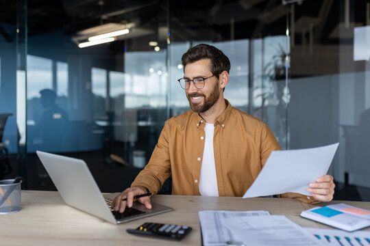 Satisfied and confident financier accountant with paper work inside the office, smiling man works with documents and bills, successful businessman compares statistical data uses laptop, calculator.