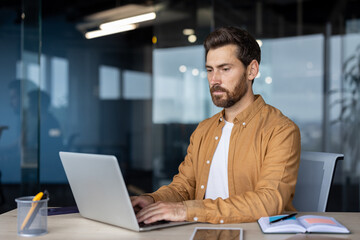 Serious man at work with laptop inside office, thinking businessman typing on keyboard, preparing electronic report, mature worker programmer, coding new software at workplace.