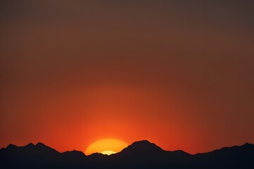 Obraz premium Silhouetted mountains at dusk with a bright orange sun dipping below the horizon, AI Generated