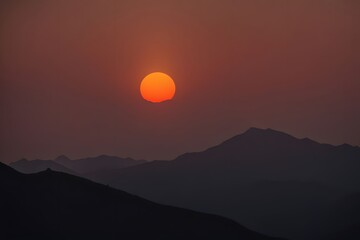 Silhouetted mountains at dusk with a bright orange sun dipping below the horizon, AI Generated