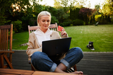 Mature woman enjoying afternoon sitting on garden chair with laptop eating watermelon in backyard