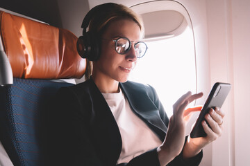 Young caucasian woman sitting near window with smartphone connected to wifi internet during flight...