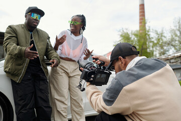 Two individuals engaging in animated conversation while being filmed outdoors by a videographer....