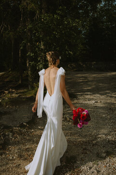 A modern bride in a white, open-back wedding dress walks along a gravel path, holding a vibrant bouquet of red and pink flowers, surrounded by natural woodland scenery.