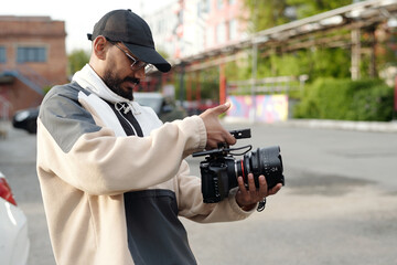 Man wearing cap handling professional camera in outdoor setting near building. Capturing video...
