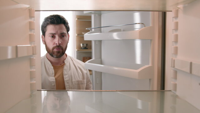 Hungry Caucasian bachelor man looking for food in empty fridge finding no meal products feeling sad disappointed poor unemployment need delivery service POV point of view from inside refrigerator