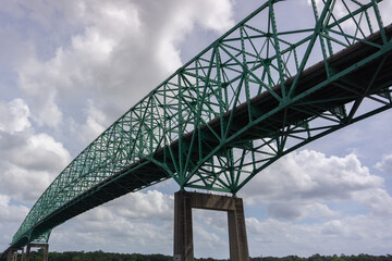 A depiction of a truss bridge against a bright blue sky, as viewed from the river beneath.