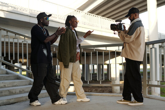 Three individuals filming under an architectural structure holding professional camera. One person is holding camera while two others gesturing and engaging in discussion