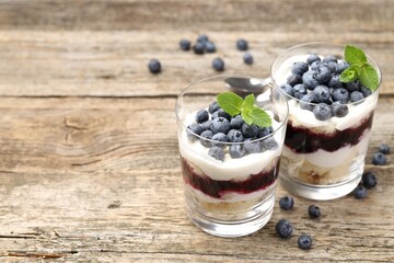 Tasty trifle dessert. Sponge cake, blueberries, jam and whipped cream in glasses on wooden table, closeup. Space for text