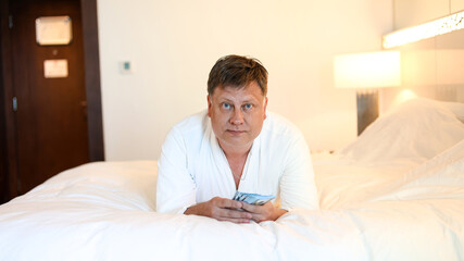 Obraz premium Man in white robe lying on bed counting cash dollars concept of successful life.