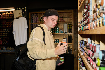 Young man with backpack looking at black can in his hand while choosing graffiti paints in front of display with stack of dispensers