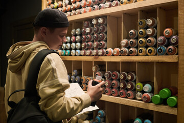Young creative graffiti artist choosing new paints while standing by display and looking at can of black color in shop