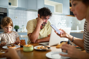 Father working on laptop while family has breakfast in kitchen