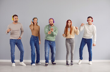 Fototapeta premium Portrait of a young joyful happy people friends or students men and women wearing casual clothes having fun listening to music in headphones standing in a row on a gray wall background.