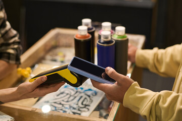 Hands of young unrecognizable male buyer of paints holding smartphone over payment terminal held by shop assistant