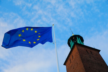 The European Union flag flutters in the breeze beside a tall brick tower of Stockholm City Hall, with a bright blue sky creating a vibrant backdrop during the day.
