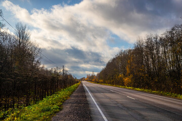A serene road framed by trees under a dramatic sky in the early hours of autumn, illuminated by a bright sky showing dynamic clouds, leads to the horizon.