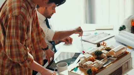 Smart architect engineer inspect house model while happy project manager walking and holding blueprint and asking about building construction. Group of diverse engineer working together. Alimentation.
