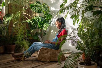 Calm smiling black woman works on laptop among plants, managing small flower business. African...