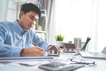 A man is sitting at a desk with a house model and a calculator. He is writing on a piece of paper