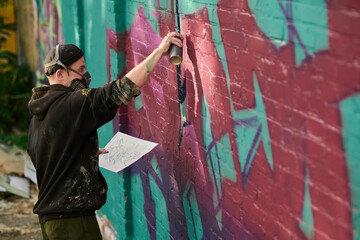 Young creative man with sketch on paper applying pink paint on brick wall while working over new graffiti masterpiece outdoors