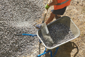 Builder fills a wheelbarrow with gravel using a shovel, working with gravel at a construction site,...