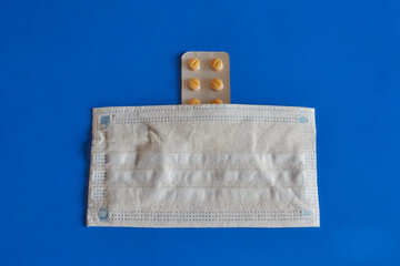 A protective medical mask lying on a blister pack of pills on a blue background, disease...