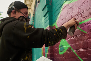 Side view of young man in hoodie and respirator looking at brick wall and spraying paint from can while creating new artwork