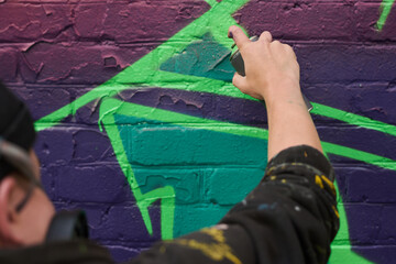 Hand of young unrecognizable man with can of graffiti paint spraying it on brick wall while outlining purple letter or figure