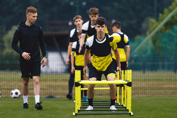 Teenagers on soccer training camp. Boys practice football with young coaches. Junior-level athletes improving soccer skills on outdoor training. The player jumping over hurdles and ladder-skipping