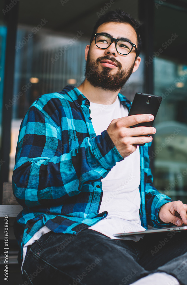 Poster kazakh young man freelancer holding smartphone gadget while working at modern laptop computer using 