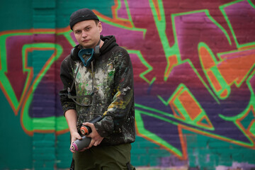 Creative and skilled young man in casual apparel holding cans with graffiti paints of orange and pink color and looking at you
