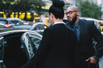 Gentle male proud ceo dressed in elegant formal wear helping female colleague sit down in car transport, African American man opening automobile car after success meeting with woman enterprise boss