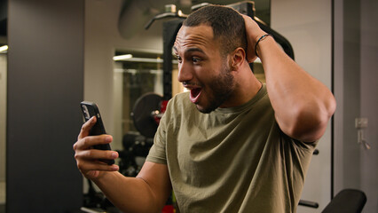 Shocked wonder surprised excited African American man trainer guy sportsman male athlete bodybuilder in sport club gym happy win victory achieve success with mobile phone message winning say wow shock