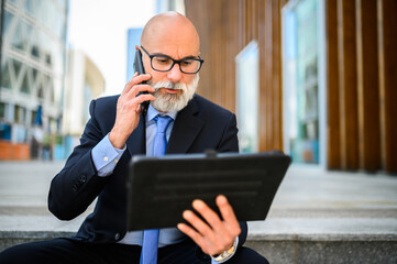 Senior businessman multitasking using phone and digital tablet outside office building