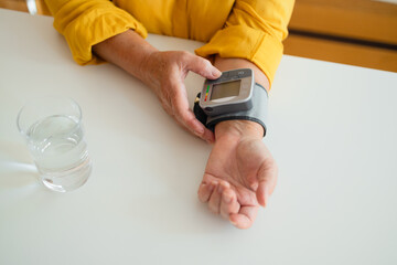 Healthy woman self checking blood pressure using machine. Senior woman with hypertension measuring blood pressure herself at home. Heart rate measurement. Glass of water on the table