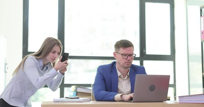 Woman employee captures photo on smartphone of male colleague in glasses. Concept of unscrupulous behavior of office workers to gain career success