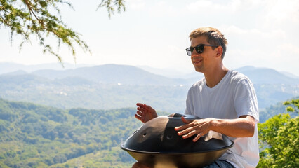 A handsome young man plays a handpan against the background of mountains.